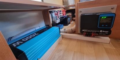 Offenes Holzfach mit links einem blauen Ger&auml;t auf dem Boden mit Aufschrift '2000W', rechts ein schwarzes Ger&auml;t mit Display und gr&uuml;nen LED-Leuchten, mehrere Kabel und Steckverbinder sichtbar