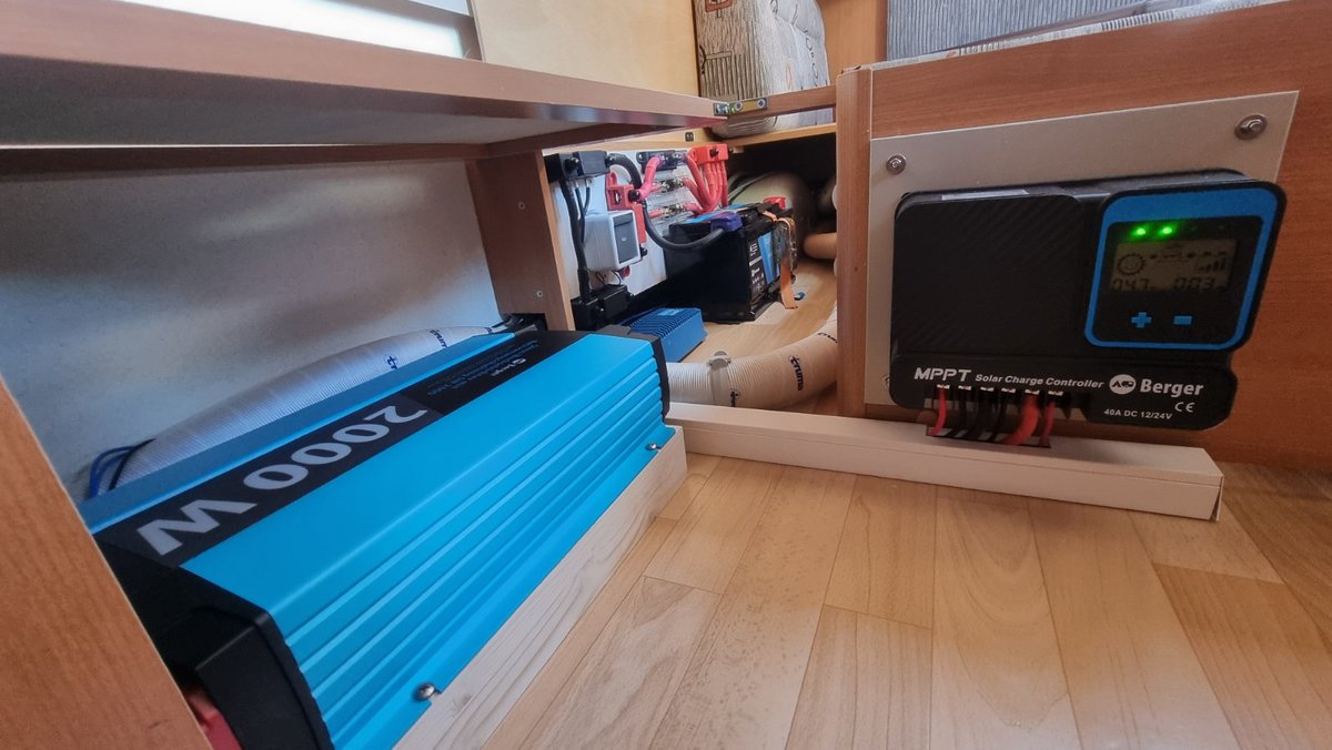 Offenes Holzfach mit links einem blauen Ger&auml;t auf dem Boden mit Aufschrift '2000W', rechts ein schwarzes Ger&auml;t mit Display und gr&uuml;nen LED-Leuchten, mehrere Kabel und Steckverbinder sichtbar