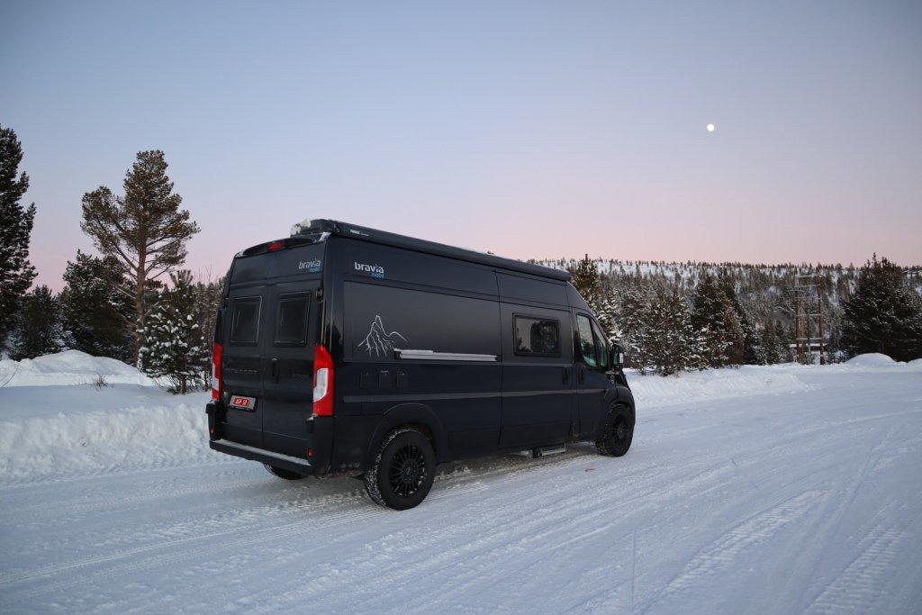 Schwarzer Camping-Van auf verschneiter Stra&szlig;e, schneebedeckte B&auml;ume im Hintergrund, heller Himmel mit sichtbarem Mond