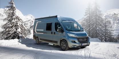Blauer Camping-Van auf verschneiter Stra&szlig;e, Tannen und Berge im Hintergrund