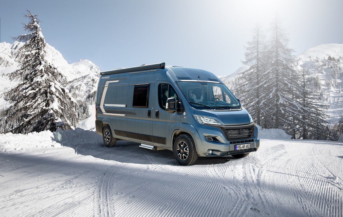 Blauer Camping-Van auf verschneiter Stra&szlig;e, Tannen und Berge im Hintergrund