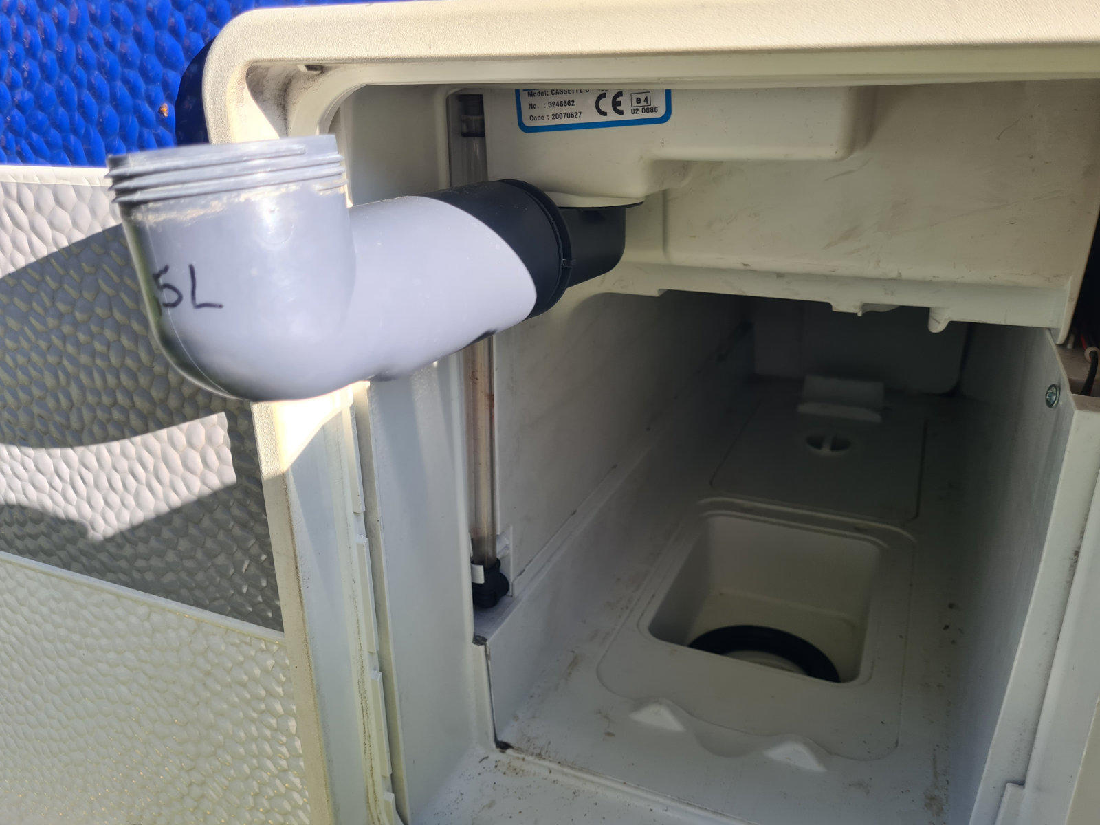 Offenes wei&szlig;es Kunststofffach mit rechteckigem Innenraum und rundem Auslass am Boden. Oben links ein grauer gebogener Rohrabschnitt mit schwarzem Verbindungsst&uuml;ck und handschriftlicher Markierung. Links au&szlig;en eine blaue strukturierte Fl&auml;che