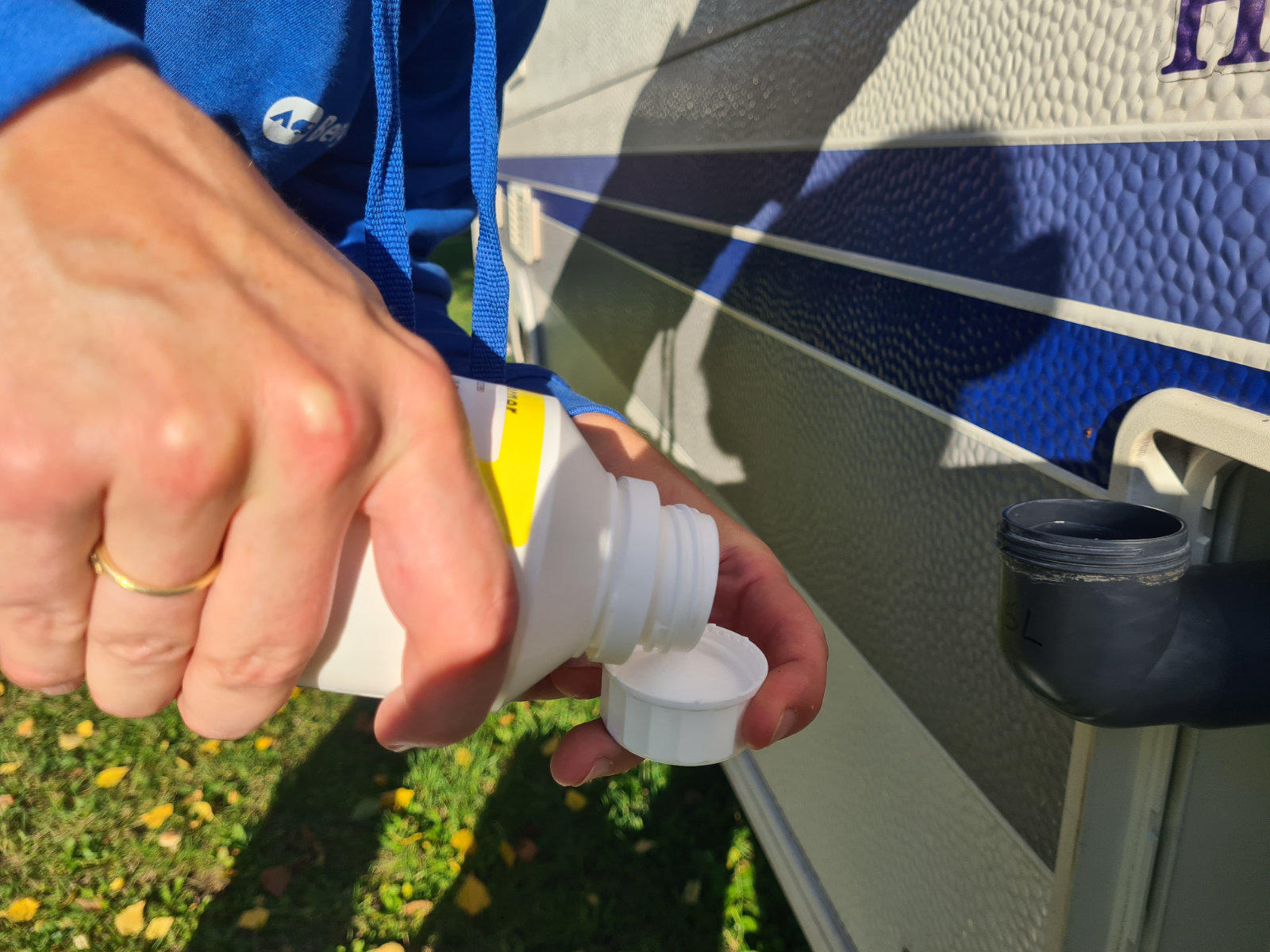 Zwei H&auml;nde halten eine wei&szlig;e Plastikflasche und gie&szlig;en Fl&uuml;ssigkeit in einen wei&szlig;en Verschluss an der Seite eines Wohnwagens