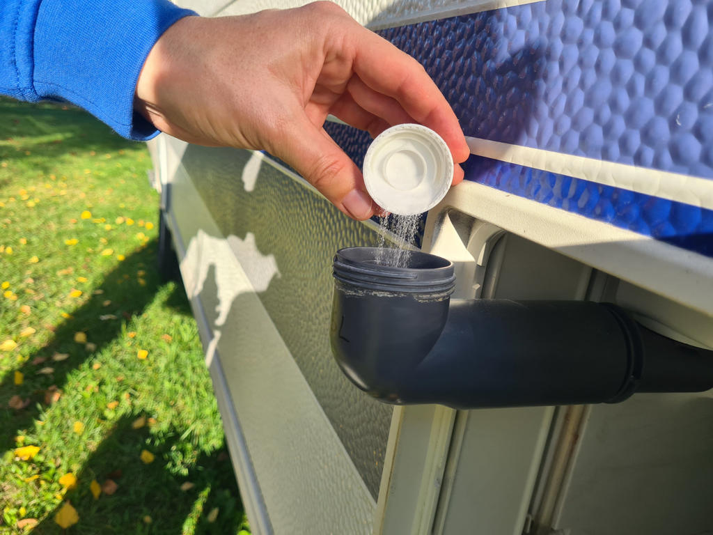 Hand sch&uuml;ttet wei&szlig;es Pulver aus einer kleinen Kunststoffkappe in ein schwarzes Ablassrohr an der Seite eines blauen Aufstellpools; Gras im Hintergrund