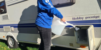 Person in blauem Kapuzenpullover gie&szlig;t Wasser aus einem wei&szlig;en Kanister in ein Fach an der Au&szlig;enseite eines Wohnwagens mit Aufschrift "Hobby prestige"; Gras und Rad sichtbar