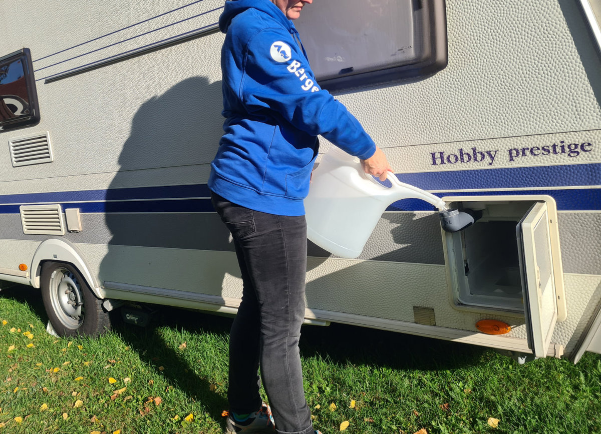 Person in blauem Kapuzenpullover gie&szlig;t Wasser aus einem wei&szlig;en Kanister in ein Fach an der Au&szlig;enseite eines Wohnwagens mit Aufschrift "Hobby prestige"; Gras und Rad sichtbar