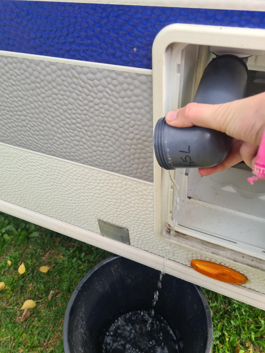Hand h&auml;lt einen grauen Ablaufstutzen aus einer offenen Klappe an der Seitenwand eines Wohnwagens; Wasser flie&szlig;t in einen schwarzen Eimer auf Gras; blaue und graue Paneele und ein orangefarbener Reflektor sichtbar