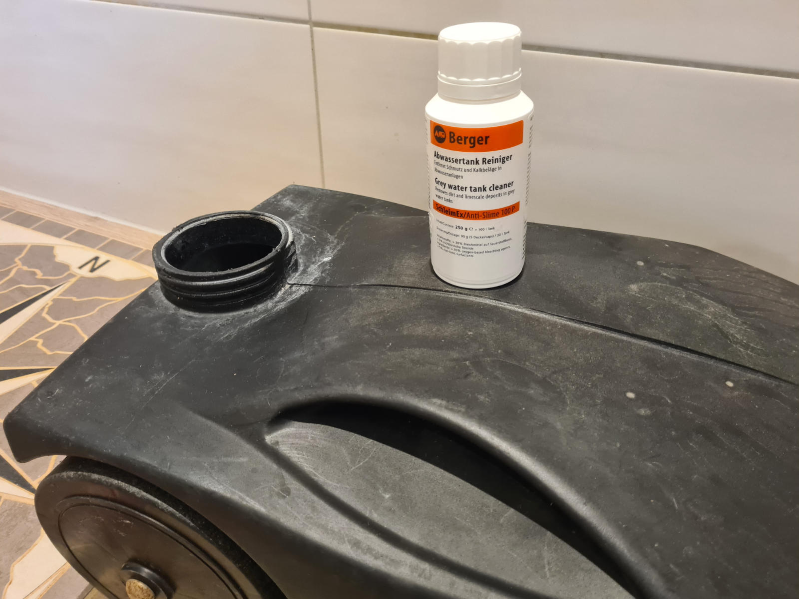 Schwarzer Kunststoffbeh&auml;lter mit offener runder &Ouml;ffnung, wei&szlig;e Flasche mit orangefarbenem Etikett darauf, geflieste Wand im Hintergrund, sichtbares Rad an der Seite