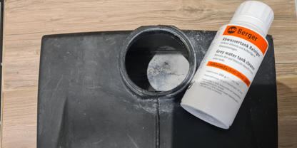 Schwarzer Kunststoffbeh&auml;lter mit runder &Ouml;ffnung; wei&szlig;e Flasche mit orangefarbenem Etikett liegt auf der Kante; heller Holzfu&szlig;boden sichtbar