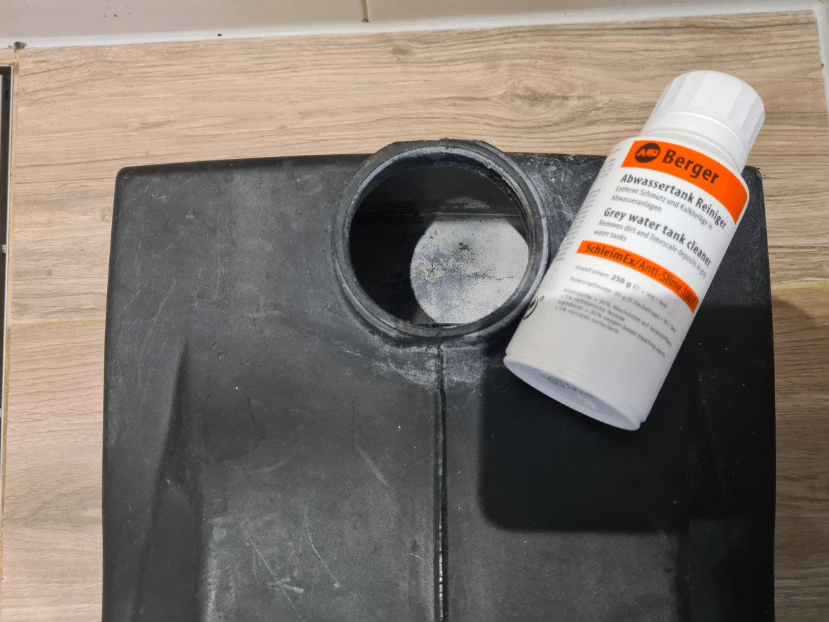 Schwarzer Kunststoffbeh&auml;lter mit runder &Ouml;ffnung; wei&szlig;e Flasche mit orangefarbenem Etikett liegt auf der Kante; heller Holzfu&szlig;boden sichtbar