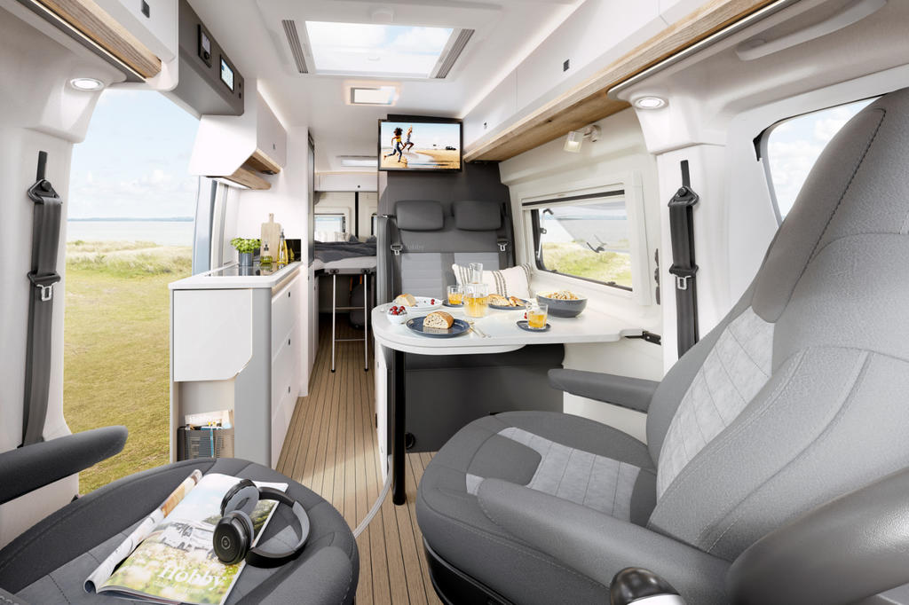 Innenraum eines Wohnmobils mit Sitzgruppe und zwei gedrehten Vordersitzen, Esstisch mit Tellern, Gläsern und Brot, Küchenzeile links, Fernseher an der Rückwand und Fenster mit Strandblick
