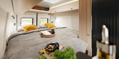 Doppelbett mit grauer Bettdecke und gelben Kissen in einem Wohnmobil, Fenster und Dachluke, Holzschr&auml;nke, Tablett mit Handtuch, Kopfh&ouml;rer und kleine gr&uuml;ne Pflanze im Vordergrund