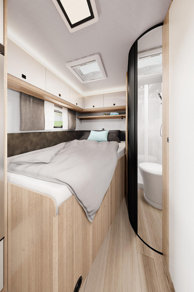 Kompaktes Schlafabteil mit Doppelbett, zwei Kissen, Decke, Holzschränken, Dachluke, Seitenfenster, schmalem Gang und sichtbarer Toilette hinter einer gebogenen Tür