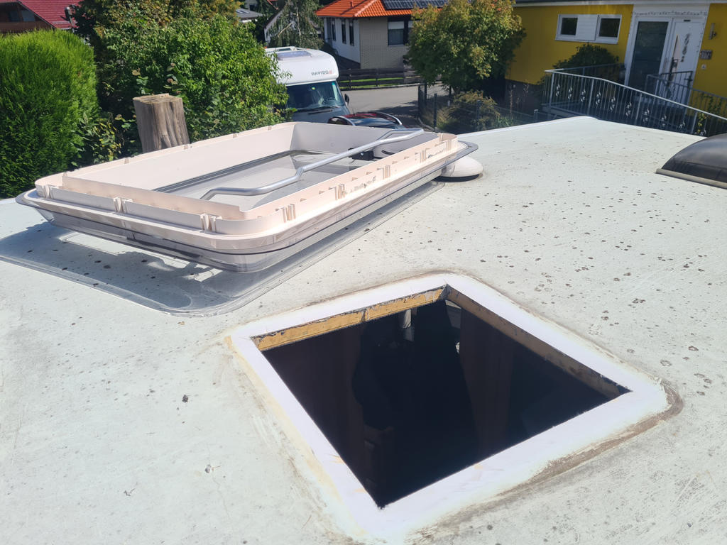 Weißes flaches Dach mit zwei rechteckigen Dachluken, eine Öffnung ohne Abdeckung und eine mit Haube; im Hintergrund Häuser, ein Lieferwagen und Bäume