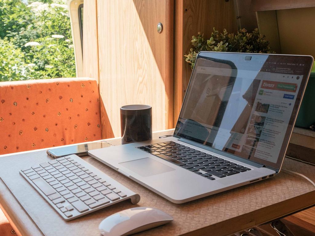 Auf einem Holztisch ein aufgeklappter Laptop, eine kabellose Tastatur, eine Maus und ein schwarzer zylindrischer Lautsprecher; im Hintergrund ein orange gepolsterter Sitz, Pflanzen und ein Fenster mit gr&uuml;nen B&auml;umen