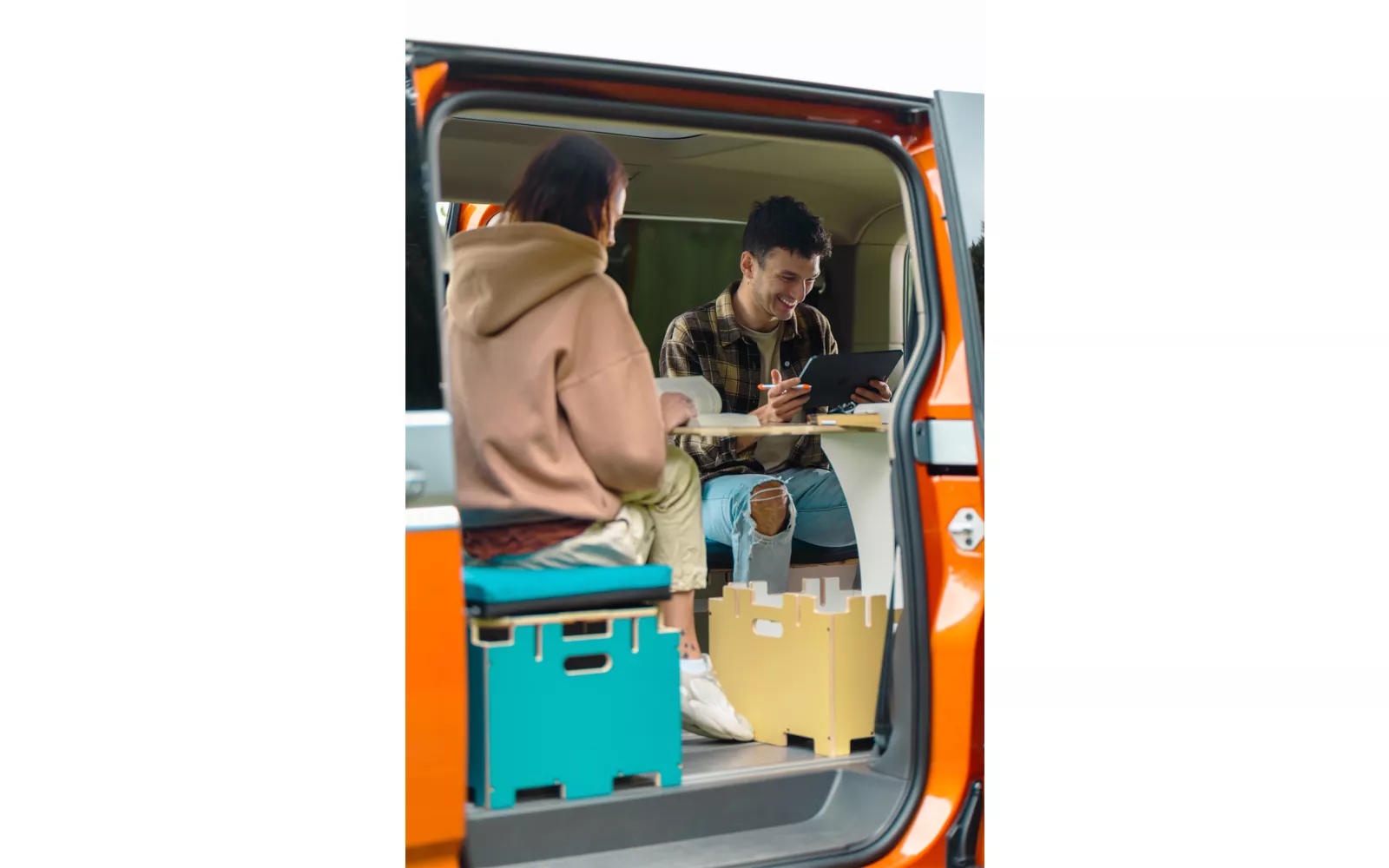R&uuml;ckseite eines orangefarbenen Vans mit offener Seitent&uuml;r, zwei Personen sitzen an einem kleinen Tisch, eine Person h&auml;lt einen Stift, bunte Aufbewahrungsboxen auf dem Fahrzeugboden