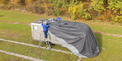 Wohnwagen auf Grasfl&auml;che, teils mit gro&szlig;er schwarzer Plane bedeckt; zwei Personen in blauen Jacken auf Leitern neben dem Wagen; im Hintergrund B&auml;ume mit gelben und orangenen Bl&auml;ttern