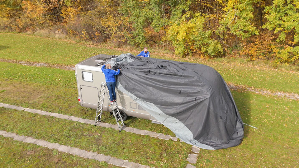 Wohnwagen auf Grasfl&auml;che, teils mit gro&szlig;er schwarzer Plane bedeckt; zwei Personen in blauen Jacken auf Leitern neben dem Wagen; im Hintergrund B&auml;ume mit gelben und orangenen Bl&auml;ttern