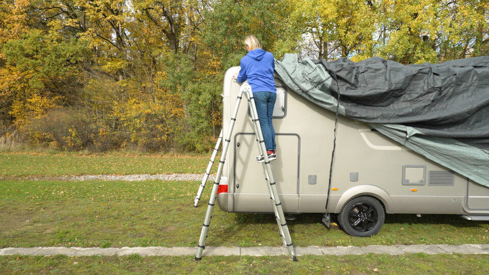 Person in blauer Jacke steht auf einer Teleskopleiter neben einem Wohnmobil; eine Plane liegt teilweise auf dem Dach; herbstliche B&auml;ume im Hintergrund, Gras und ein Kiesweg im Vordergrund