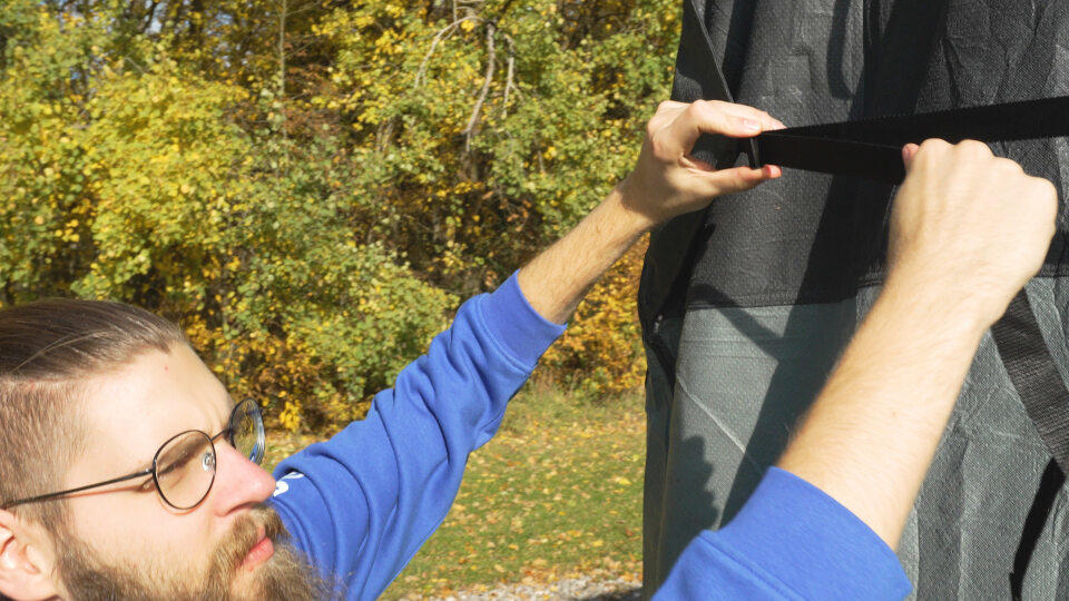 Mann mit Brille und Bart zieht schwarzen Stoff an einen grauen Rahmen, im Hintergrund B&auml;ume mit Herbstlaub