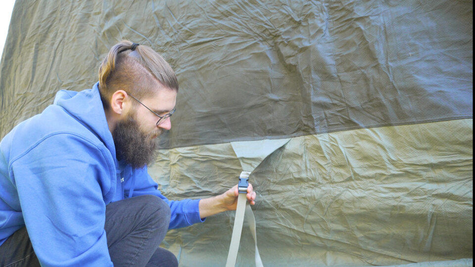 Person mit Bart, Brille und blauem Kapuzenpullover kniet und h&auml;lt einen Riemen an einem gr&uuml;nen Zelt