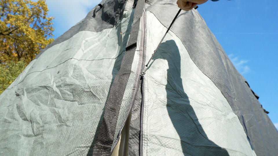 Zeltstoff mit mittigem Rei&szlig;verschluss und Netz, Schatten einer Person mit Hut auf dem Stoff, blauer Himmel und Baum mit gelben Bl&auml;ttern im Hintergrund