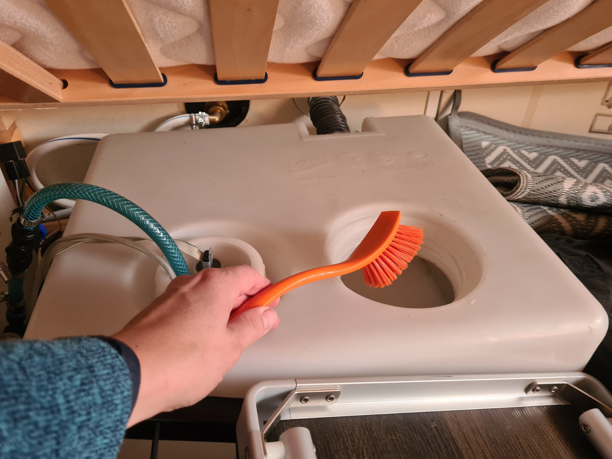 Hand h&auml;lt orangefarbene B&uuml;rste &uuml;ber hellgra&szlig;en Kunststofftank mit zwei runden &Ouml;ffnungen, gr&uuml;nem Schlauch und Holzlatten dar&uuml;ber