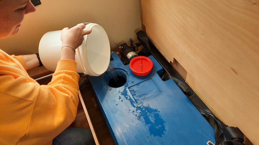 Person gie&szlig;t Wasser aus einer wei&szlig;en Sch&uuml;ssel in einen blauen Plastikbeh&auml;lter mit runden &Ouml;ffnungen und roter Schraubkappe in einem Holzfach