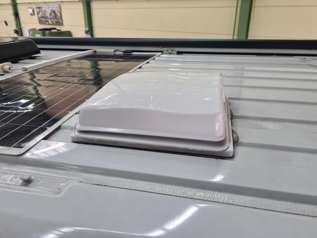 Wei&szlig;e rechteckige Kunststoffabdeckung auf grauem, profiliertem Metalldach neben einem Solarpanel; gr&uuml;ne Stahltr&auml;ger und Wand im Hintergrund