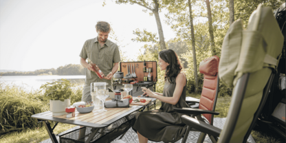Ein Mann gie&szlig;t ein Getr&auml;nk an einem Klapptisch, eine Frau sitzt daneben; auf dem Tisch Tassen, Teller, Obstschale und Kaffeekanne; zwei gro&szlig;e St&uuml;hle, ein Rucksack, See und B&auml;ume im Hintergrund