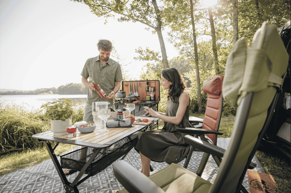 Ein Mann gie&szlig;t ein Getr&auml;nk an einem Klapptisch, eine Frau sitzt daneben; auf dem Tisch Tassen, Teller, Obstschale und Kaffeekanne; zwei gro&szlig;e St&uuml;hle, ein Rucksack, See und B&auml;ume im Hintergrund