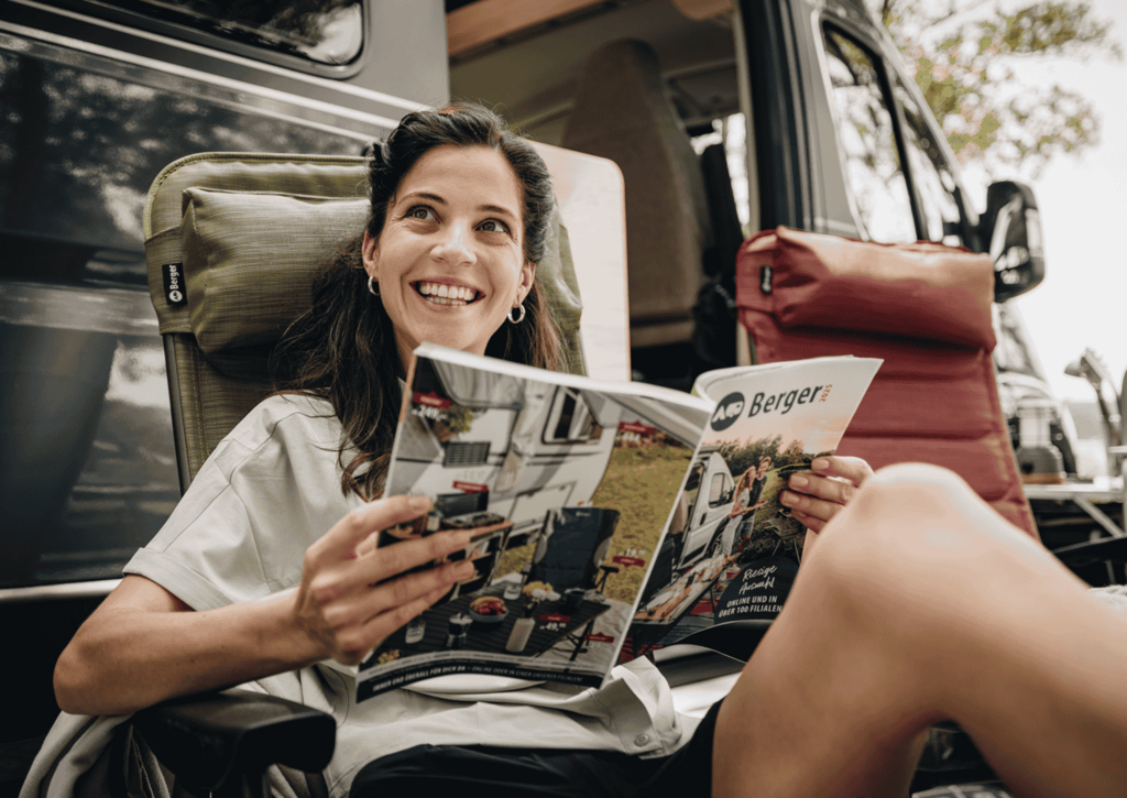 Frau auf Campingstuhl vor Kastenwagen mit Zeitung in der Hand