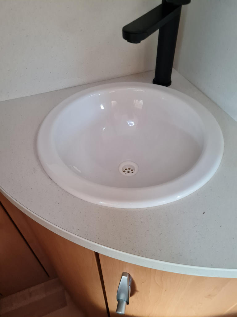 Round white sink in a light countertop, black faucet, wooden cabinet with silver handle below