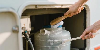 Zwei H&auml;nde reinigen eine graue Metallflasche in einem offenen Staufach eines Wohnwagens mit einer B&uuml;rste und einem flachen Werkzeug; Seifenschaum auf der Flasche