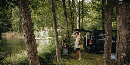 Person zieht T‑Shirt &uuml;ber den Kopf neben offenem schwarzem Van am See, umgeben von Birken