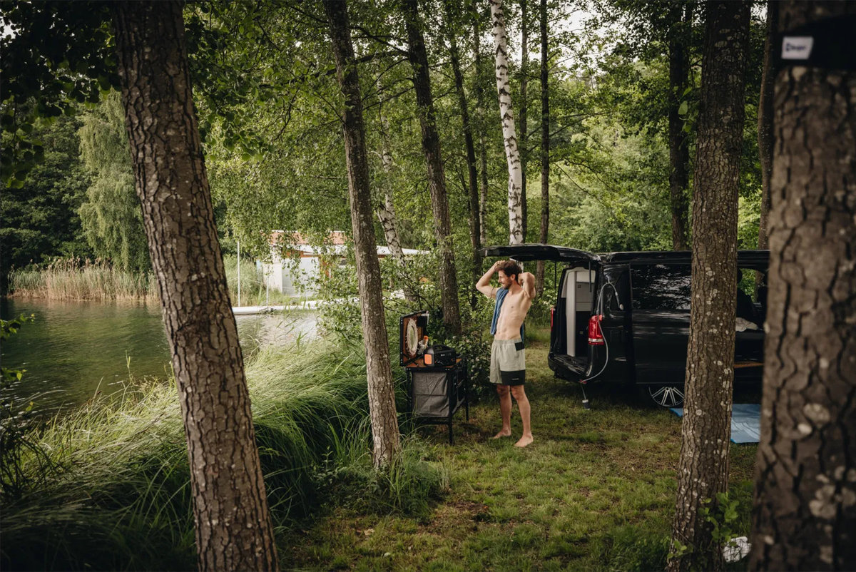 Person zieht T‑Shirt &uuml;ber den Kopf neben offenem schwarzem Van am See, umgeben von Birken