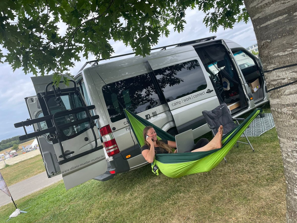 Person in einer gr&uuml;nen H&auml;ngematte, die zwischen einem Baum und einem silbernen Wohnmobil befestigt ist; die Person h&auml;lt einen wei&szlig;en Becher an den Mund, ein aufgeklappter Laptop liegt auf dem Scho&szlig;; die Schiebet&uuml;r des Wohnmobils ist offen; Fahrzeug s