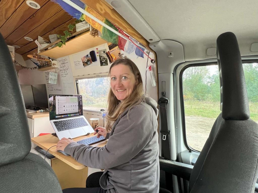 Frau sitzt an einem Holztisch im Camper, blickt in die Kamera, offenes Laptop auf dem Tisch, Papiere und Stift, Fenster mit Au&szlig;enblick, Regal mit Pflanzen und Fotos