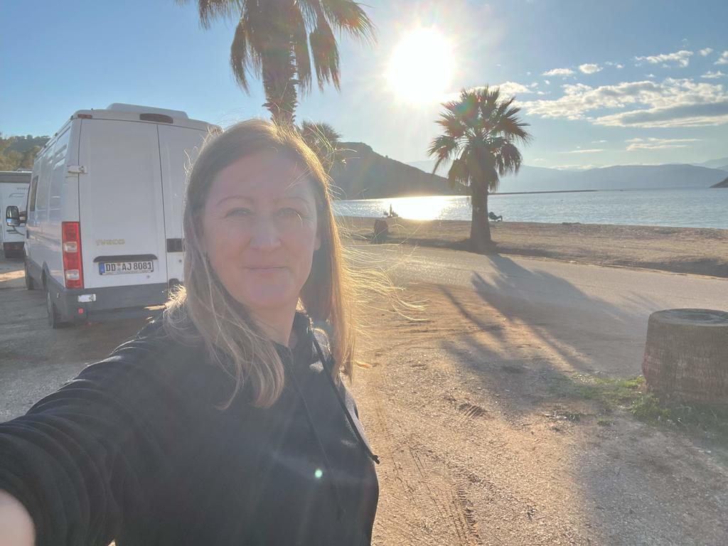 Person mit langen blonden Haaren im Vordergrund, Arm ausgestreckt zur Kamera. Strand mit Palmen, Meer, tiefer stehende Sonne am Himmel und ein geparkter wei&szlig;er Van im Hintergrund