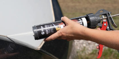 Zwei H&auml;nde halten eine Kartuschenpresse mit schwarzer Kartusche; die Kartuschenspitze steht an der Kante eines hellgrauen Autoteils