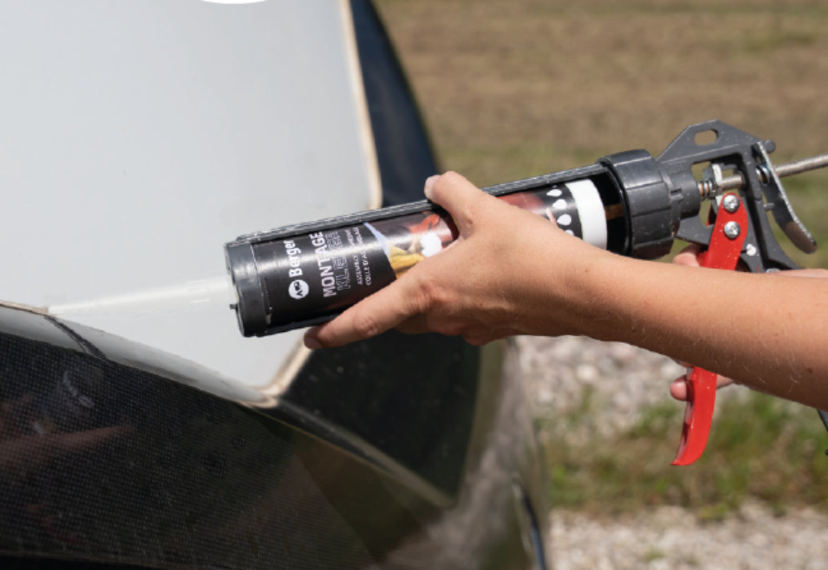 Zwei H&auml;nde halten eine Kartuschenpresse mit schwarzer Kartusche; die Kartuschenspitze steht an der Kante eines hellgrauen Autoteils