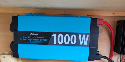 Blau-schwarzes rechteckiges Ger&auml;t mit Aufdruck "1000 W", zwei angeschlossene Kabel (rot und schwarz), auf einer Holzplatte montiert; links ein beige-zylindrisches Objekt, rechts ein teilweise sichtbares schwarzes Ger&auml;t