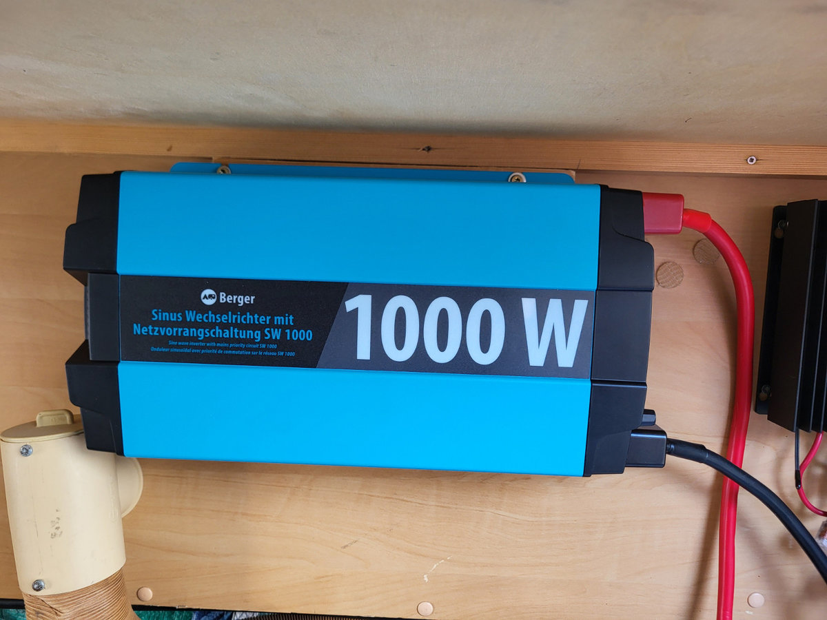 Blau-schwarzes rechteckiges Ger&auml;t mit Aufdruck "1000 W", zwei angeschlossene Kabel (rot und schwarz), auf einer Holzplatte montiert; links ein beige-zylindrisches Objekt, rechts ein teilweise sichtbares schwarzes Ger&auml;t