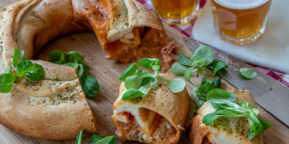 Aufgeschnittenes, ringf&ouml;rmiges gef&uuml;lltes Brot mit geschmolzenem K&auml;se und roter F&uuml;llung auf einem Holzbrett, gr&uuml;ne Bl&auml;tter, ein Messer und zwei Gl&auml;ser helles Bier im Hintergrund