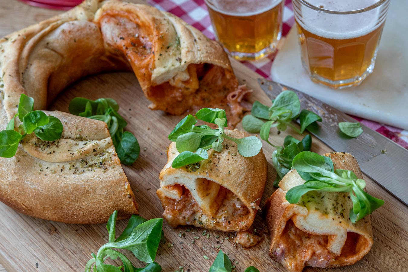Aufgeschnittenes, ringf&ouml;rmiges gef&uuml;lltes Brot mit geschmolzenem K&auml;se und roter F&uuml;llung auf einem Holzbrett, gr&uuml;ne Bl&auml;tter, ein Messer und zwei Gl&auml;ser helles Bier im Hintergrund