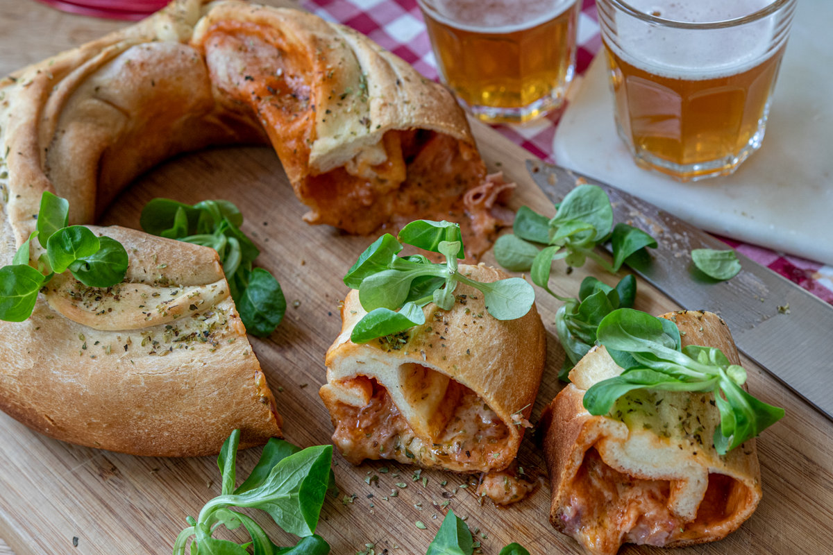 Aufgeschnittenes, ringf&ouml;rmiges gef&uuml;lltes Brot mit geschmolzenem K&auml;se und roter F&uuml;llung auf einem Holzbrett, gr&uuml;ne Bl&auml;tter, ein Messer und zwei Gl&auml;ser helles Bier im Hintergrund