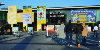 Vier Personen von hinten gehen auf einen gro&szlig;en Eingang mit den Schildern 'Eingang Ost' und 'Entrance East'. Bunte Werbebanner an der Fassade, blauer Himmel