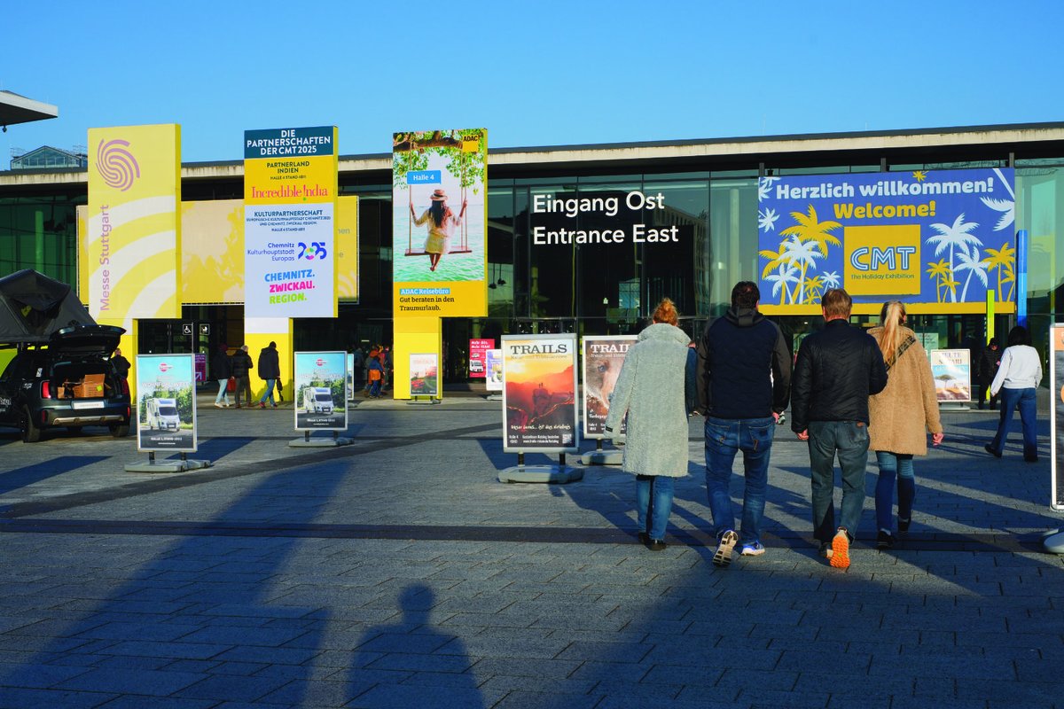 Vier Personen von hinten gehen auf einen gro&szlig;en Eingang mit den Schildern 'Eingang Ost' und 'Entrance East'. Bunte Werbebanner an der Fassade, blauer Himmel