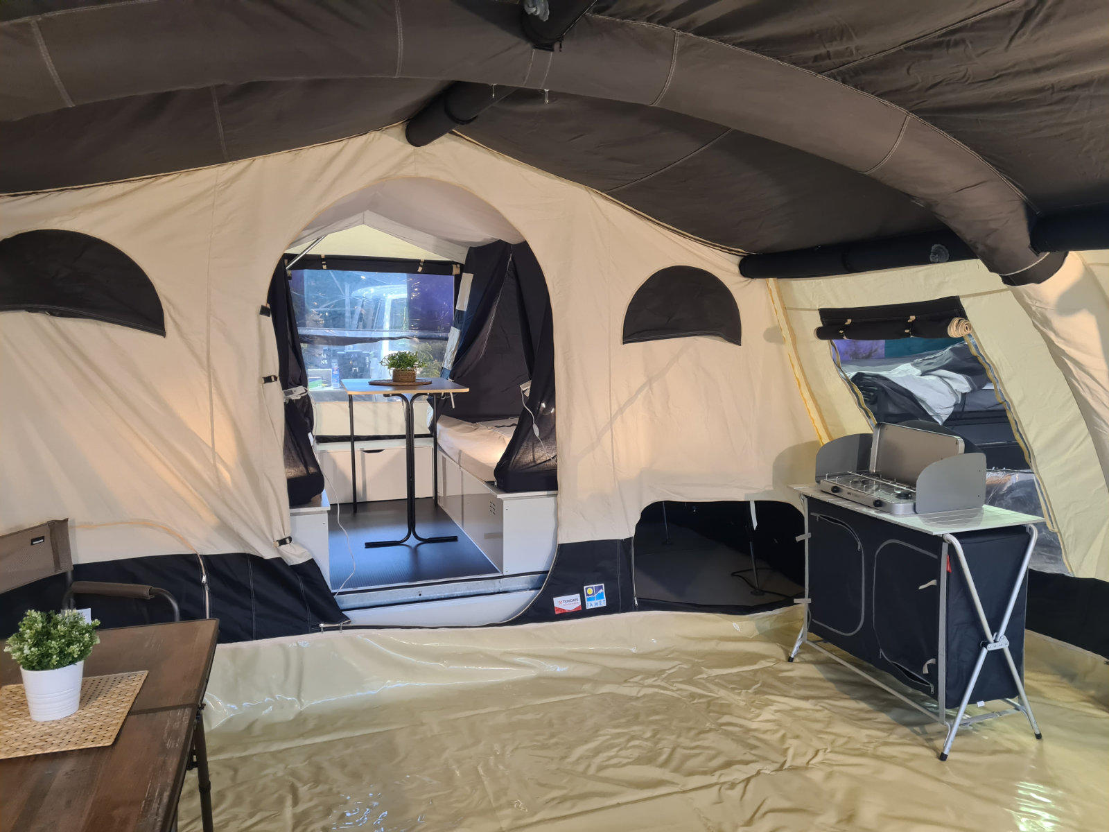 Innenraum eines beigen Zeltes mit dunkelgrauen Dachst&uuml;tzen, Bodenplane, links ein Holztisch mit Topfpflanze, rechts ein grauer K&uuml;chenblock mit Arbeitsplatte und F&auml;chern; durch einen offenen Bogen runder Tisch und zwei St&uuml;hle