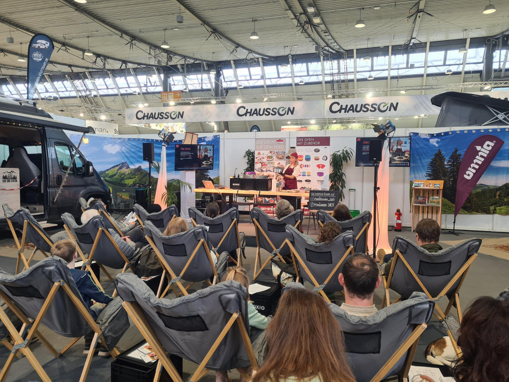 Reihen grauer faltbarer Liegest&uuml;hle mit sitzenden Personen vor einer kleinen B&uuml;hne. Auf der B&uuml;hne zwei Personen an einem Tisch, gro&szlig;e 'CHAUSSON'-Schriftz&uuml;ge an der R&uuml;ckwand, links ein Wohnmobil, hohe Hallendecke mit Dachfenstern